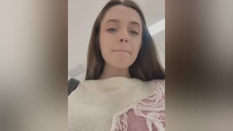 Media: A video of a young woman with fair skin and long brown hair, wearing a cream-colored sweater with pink fringes, taken from a slightly low angle, creating a sense of intimacy.