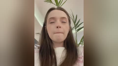 Media: Video of a young Caucasian woman with long brown hair, wearing a white sweater, with her eyes closed and a neutral expression, surrounded by green houseplants.