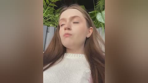 Media: Video of a young woman with fair skin, long brown hair, and closed eyes, wearing a white knit sweater, standing amidst lush green foliage.