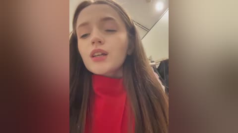 Media: Video of a young woman with long brown hair, wearing a red turtleneck, leaning forward with eyes closed, in a dimly lit room.