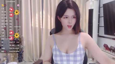 Media: A video of a fair-skinned East Asian woman with straight black hair, wearing a white checkered tank top, sitting at a desk. Background includes a calendar, sunflowers, and a television.