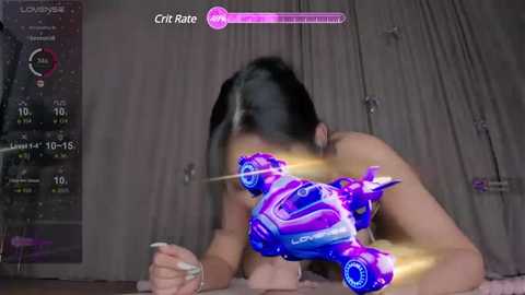 Video of a naked Asian woman with black hair, lying on a bed with a purple and blue virtual car projected over her body. Background includes a digital health monitor and curtains.