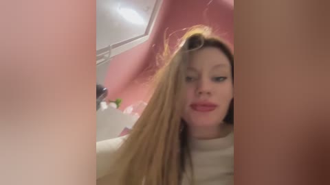 A blurry video of a young woman with long, blonde hair, wearing a white shirt, taken in a pink bathroom with a white sink and mirror in the background.