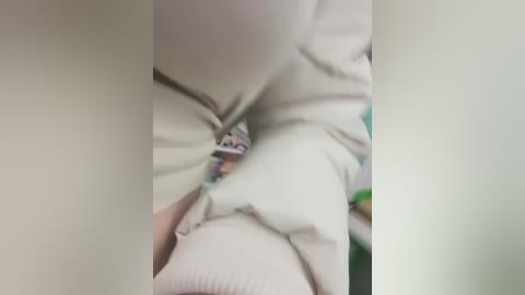 Media: Video of a person's lower torso, partially covered in light beige fabric. The fabric is crumpled, revealing a glimpse of a colorful pattern underneath. The setting appears to be indoors, with blurred background objects.