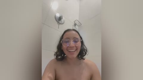 A video of a smiling, topless woman with medium skin tone and dark curly hair, wearing glasses, standing in a white-tiled shower.