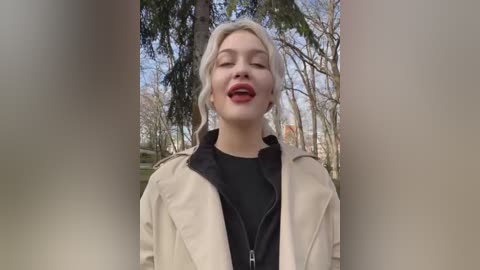 Video of a blonde woman with fair skin, wearing a beige trench coat over a black dress, smiling with red lipstick, standing outdoors in a park with bare trees and a blue sky.