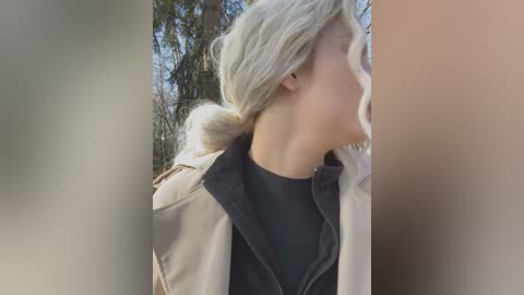 A video captures a close-up of a woman with long, platinum blonde hair tied in a low ponytail, wearing a beige jacket over a black shirt, outdoors with blurred trees in the background.