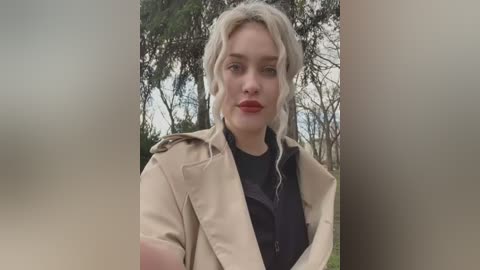 Video of a blonde woman with fair skin and red lipstick, wearing a beige trench coat over a black turtleneck, standing in a forested area with blurred trees in the background.