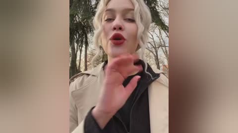 A video of a fair-skinned woman with blonde, wavy hair, wearing a beige jacket, standing in a forest. She is making a peace sign with her right hand, her lips slightly parted.