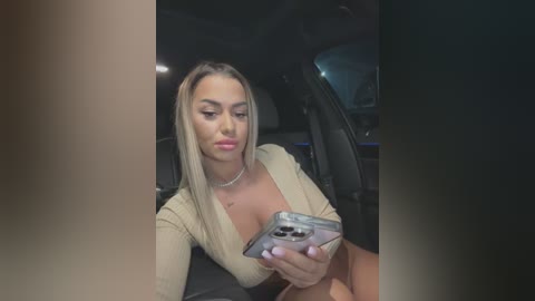 Video of a young woman with long blonde hair, wearing a beige top, taking a selfie in a car at night.