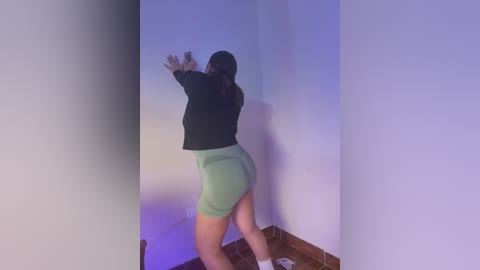 Media: Video of a woman in a green skirt and black top, reaching up against a white wall in a dimly lit room with purple lighting, standing on a wooden floor.