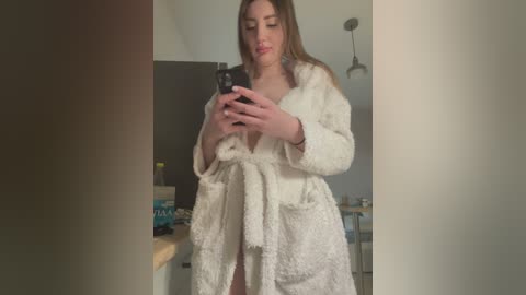 Media: Video of a young woman with long brown hair, fair skin, and a slender physique, wearing a white, plush bathrobe, taking a selfie in a modern bathroom.