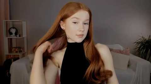 Media: Video of a fair-skinned, red-haired young woman with long hair, wearing a black halter top, adjusting her hair. Background features a white sofa, bookshelf, and plant in a softly lit room.