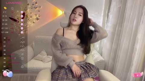 Media: Video of an Asian woman with long black hair, wearing a gray off-shoulder sweater and plaid skirt, sitting in a modern living room with white furniture and soft lighting.