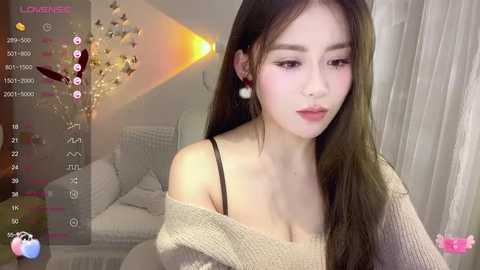 Media: Video of an East Asian woman with long, straight dark hair, fair skin, wearing a beige off-shoulder sweater, sitting in a modern living room with a couch and decorative lights.