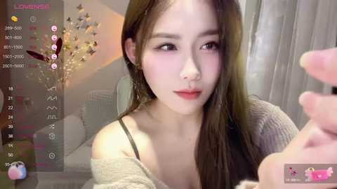 Media: A video of an East Asian woman with long brown hair and fair skin, wearing a beige off-shoulder sweater, in a cozy room with a floral arrangement and dim lighting.