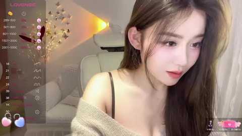 Media: A video of an East Asian woman with long, straight brown hair, wearing a beige off-shoulder sweater, in a cozy, dimly-lit room with a white couch and a glowing lamp.