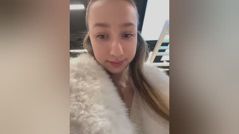 Media: A video of a young woman with light skin and straight brown hair, wearing a white fluffy jacket, smiling softly, in a modern indoor setting with black and white decor.