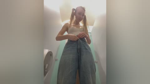 Video of a slender, blonde woman in a white tank top and high-waisted, light blue jeans, adjusting her pants, standing in a narrow, brightly lit, white-tiled bathroom with a toilet visible on the left.