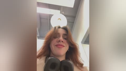 Video of a young woman with shoulder-length wavy brown hair, wearing large black headphones, smiling, in a modern, well-lit room with white ceiling and exposed beams.