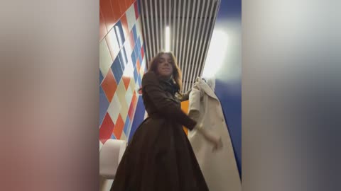 Video of a woman in a brown coat holding a white coat, standing in a restroom with bold, geometric-patterned walls.