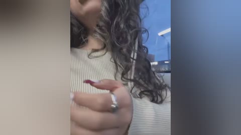 A video showing a close-up of a woman's hand holding a red nail polish bottle, with curly dark hair and a white ribbed sweater. Background features a blue wall and a white ceiling light.