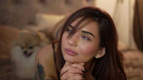 Video of a light-skinned woman with long brown hair and bangs, wearing earrings, looking contemplative, resting her chin on her hand, next to a small white and tan dog.