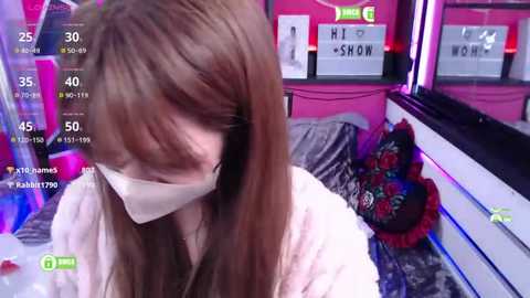 Media: Video of a young Asian woman with long brown hair and a white face mask, standing in a bright, pink-themed room with a bed covered in decorative pillows.