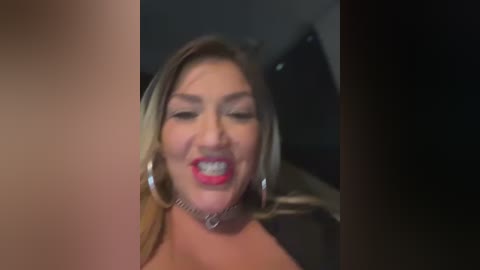 Media: A video of a Caucasian woman with light skin, long blonde hair, and red lipstick, smiling widely, wearing a silver choker and large hoop earrings. The background is blurred, possibly indoors with dark furniture.