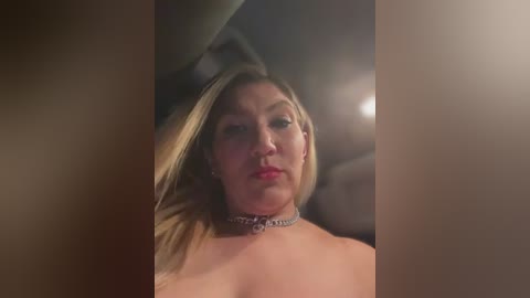 Media: A video of a topless woman with fair skin, blonde hair, and red lipstick, wearing a choker necklace, sitting in a dimly lit car.