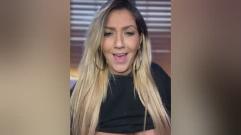 Media: A video of a woman with long, blonde ombre hair, wearing a black top, smiling with her mouth open, standing indoors with horizontal blinds in the background.