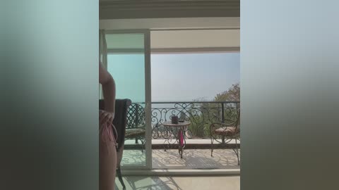 Video of a person's hand holding a pink object, peering through a frosted glass door into a sunlit balcony with a wrought-iron railing, a small table, and a distant ocean view.