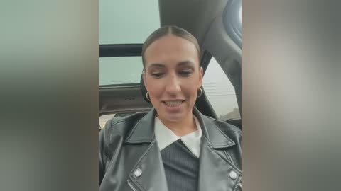 Media: Video of a woman with fair skin, brown hair tied back, wearing a black leather jacket over a white collared shirt, sitting in a car, looking amused.