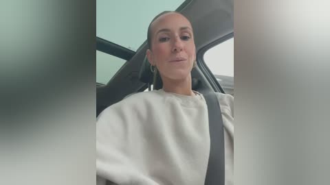 Media: Video of a Caucasian woman with a fair complexion, light skin, and short, straight, brown hair, wearing a white sweatshirt and a seatbelt. She is sitting in a car, with blurred car windows in the background.