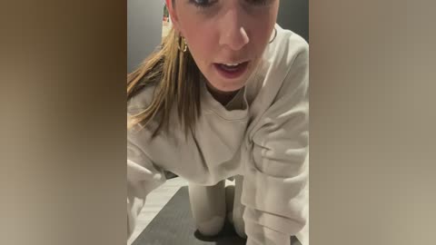 Media: Video of a woman with light skin, light brown hair tied back, wearing a beige sweatshirt, leaning over, looking upwards.