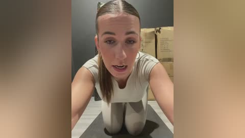 Media: A video of a Caucasian woman with fair skin, brown hair in a ponytail, wearing a white sleeveless top and light gray leggings, kneeling on a gray carpet, looking up with a surprised expression.