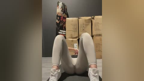 Media: Video of a woman in tight, grey leggings and white sneakers, sitting on a grey mat in a room filled with brown cardboard boxes.