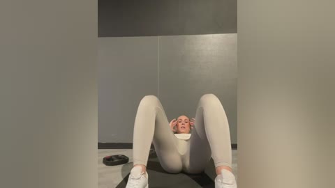 Media: Video of a blonde woman in tight white leggings and white sneakers, lying on her back on a yoga mat, legs spread wide, in a dimly lit room with grey walls.