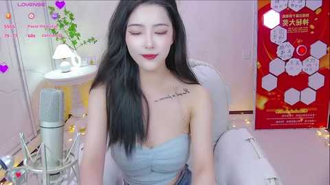 Media: Video of an Asian woman with long black hair, fair skin, and red lipstick, wearing a strapless light blue top, sitting in front of a microphone. Background features a pink wall, a plant, and a colorful poster.