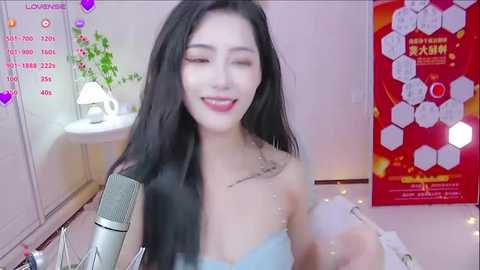 Media: Video of an East Asian woman with long black hair, fair skin, and light makeup, smiling, holding a microphone, wearing a strapless light blue top, in a softly lit, cozy bedroom with a white wall and a colorful poster.