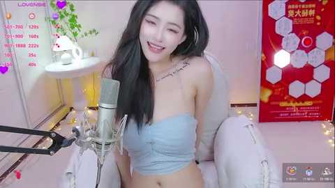 Media: A video of a smiling East Asian woman with long black hair, wearing a light blue strapless top, singing into a microphone, surrounded by a cozy, pink-themed room with hexagon-patterned posters and fairy lights.