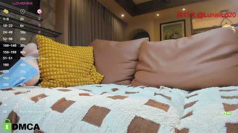 Media: A video captures a cozy bedroom with a brown leather sofa adorned with mustard-yellow pillows and a patterned blanket. The background features a warm, dimly lit room with framed art on the wall.