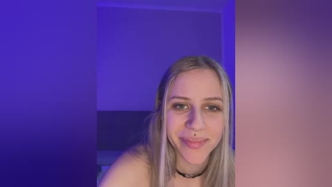 Media: Video of a young, fair-skinned, blonde woman with straight hair, wearing a black choker and a subtle smile. She stands in a dimly lit room with purple and blue lighting.