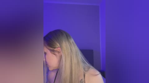 Media: Video of a young woman with long blonde hair, fair skin, and a slim figure, captured from the side, against a purple-lit wall in a dimly lit room.