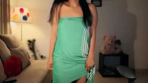 Media: Video of a woman with long black hair, wearing a strapless green dress with white stripes, standing in a dimly lit living room with a beige couch, lamp, and stuffed teddy bear.