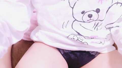 Media: Video of a person sitting with their legs apart, wearing a white sweatshirt with a cartoon bear and black lace panties.