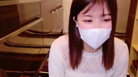 Media: Video of an Asian woman with long black hair, wearing a white face mask and white lace top, kneeling in a modern kitchen with stainless steel appliances.