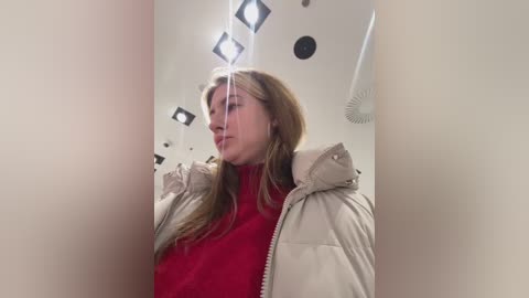 A video of a young woman with light skin and long blonde hair, wearing a red turtleneck and beige puffer jacket, standing in a modern, minimalist bathroom with white walls and square lights.