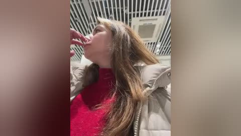 Video of a young woman with long, straight brown hair, wearing a red sweater and beige puffer jacket, licking her fingers, standing in a bathroom with a white ceiling and black grid pattern.