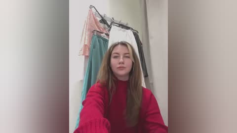 Video of a young woman with long, straight brown hair, wearing a red sweater, standing in a dimly lit room with a coat rack displaying various colorful garments.